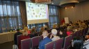 Zdjęcie przedstawia uczestników konferencji słuchających jednej z prezentacji, w tle widać osobę prezentującą oraz ekran z prezentacją kolorowych odznak.